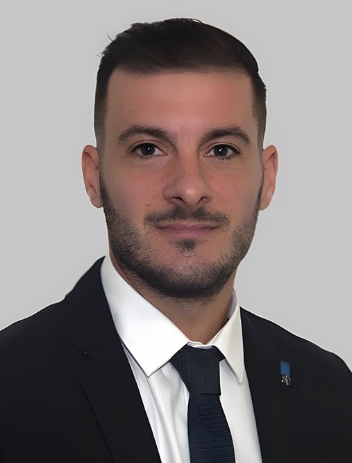 Portrait homme professionnel costume cravate homme Portrait d'un homme jeune en costume noir, chemise blanche et cravate sombre, avec une barbe soignée et une petite épinglette bleue sur le revers.